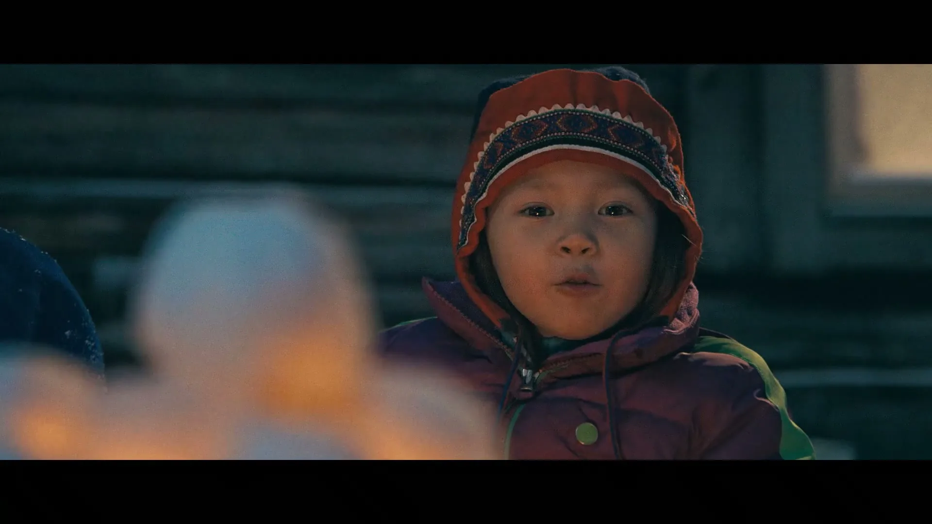 A young child in a colorful hat and winter coat looks directly at the camera, with a blurred foreground and a wooden building in the background at night.