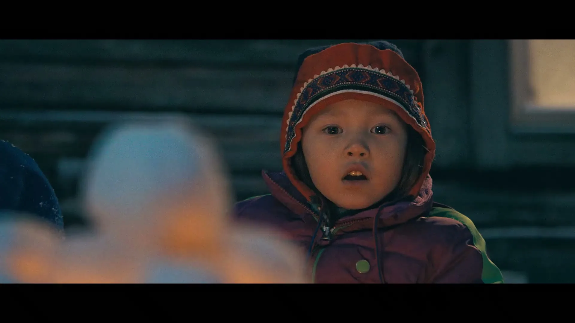 A young child in a winter hat and jacket looks intently at something off-screen, with a blurred foreground element, at night.