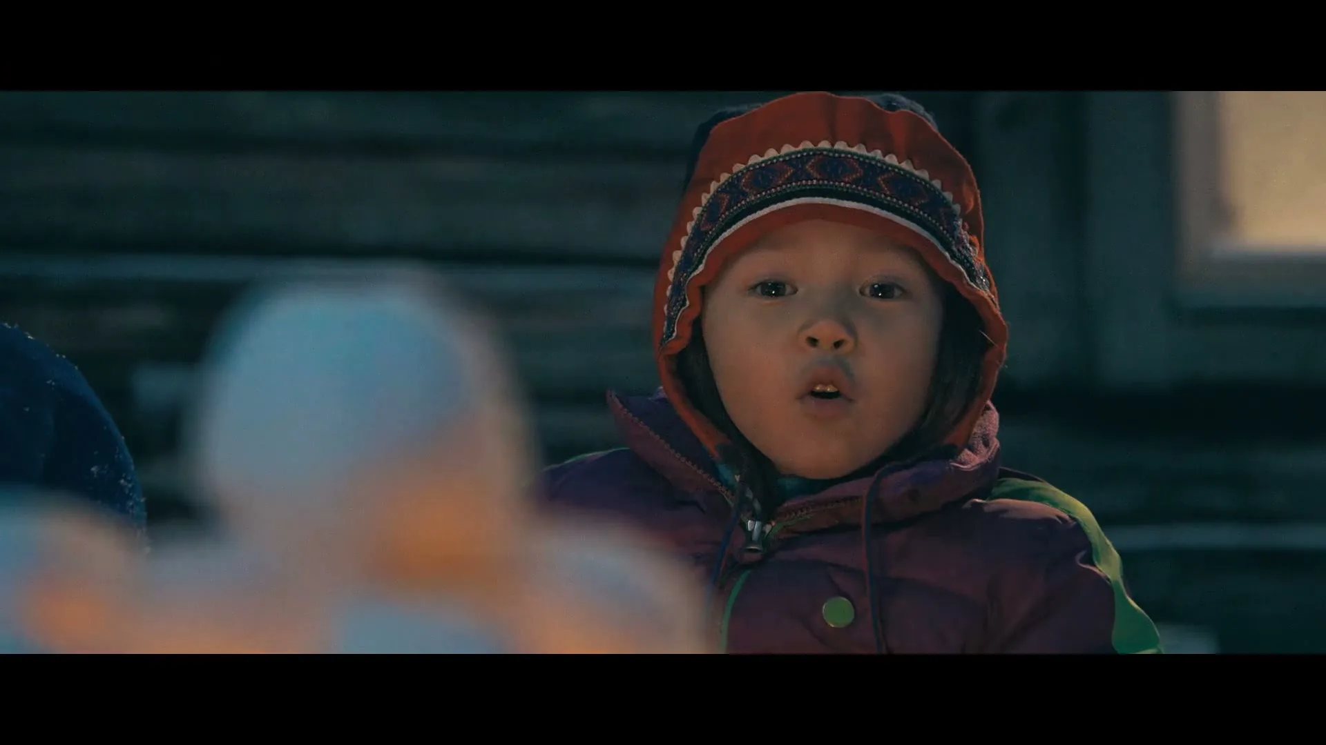 A young child in a colorful winter hat and jacket looks intently at something out of frame, with a blurred foreground element and a wooden building in the background at night.