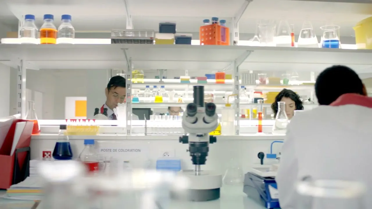 A male scientist in a lab coat works at a computer, while other scientists are visible in the background of a bright, busy laboratory.