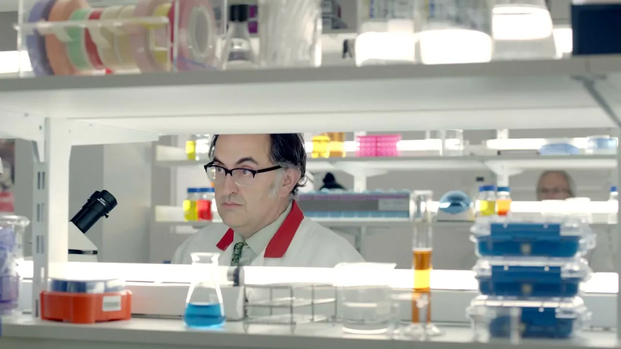 A male scientist in a lab coat and glasses looks up from his work, surrounded by lab equipment in a bright laboratory.