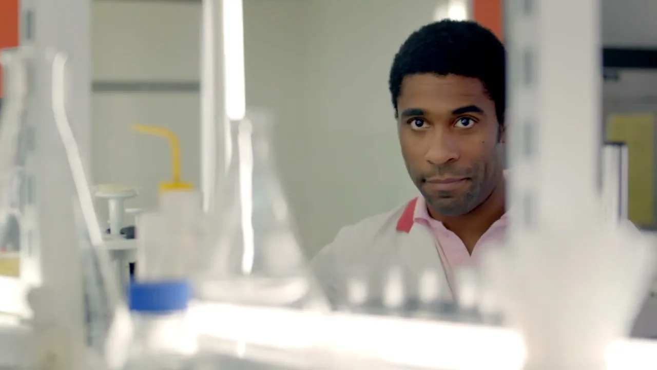 A male scientist in a lab coat peeks from behind laboratory equipment, looking directly at the camera with a curious expression in a bright lab.