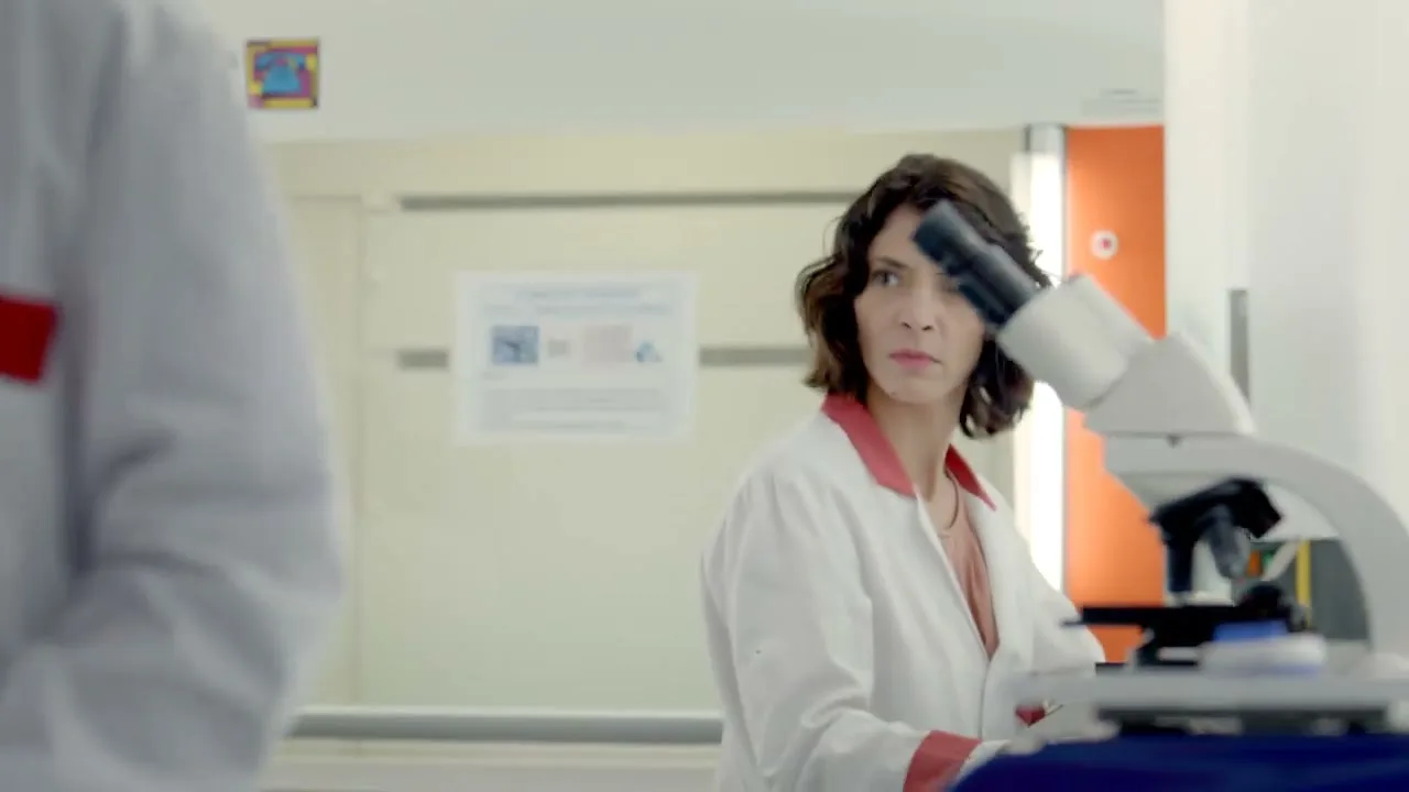 A female scientist in a lab coat looks up from a microscope with a concerned expression, as another scientist walks past in the foreground.