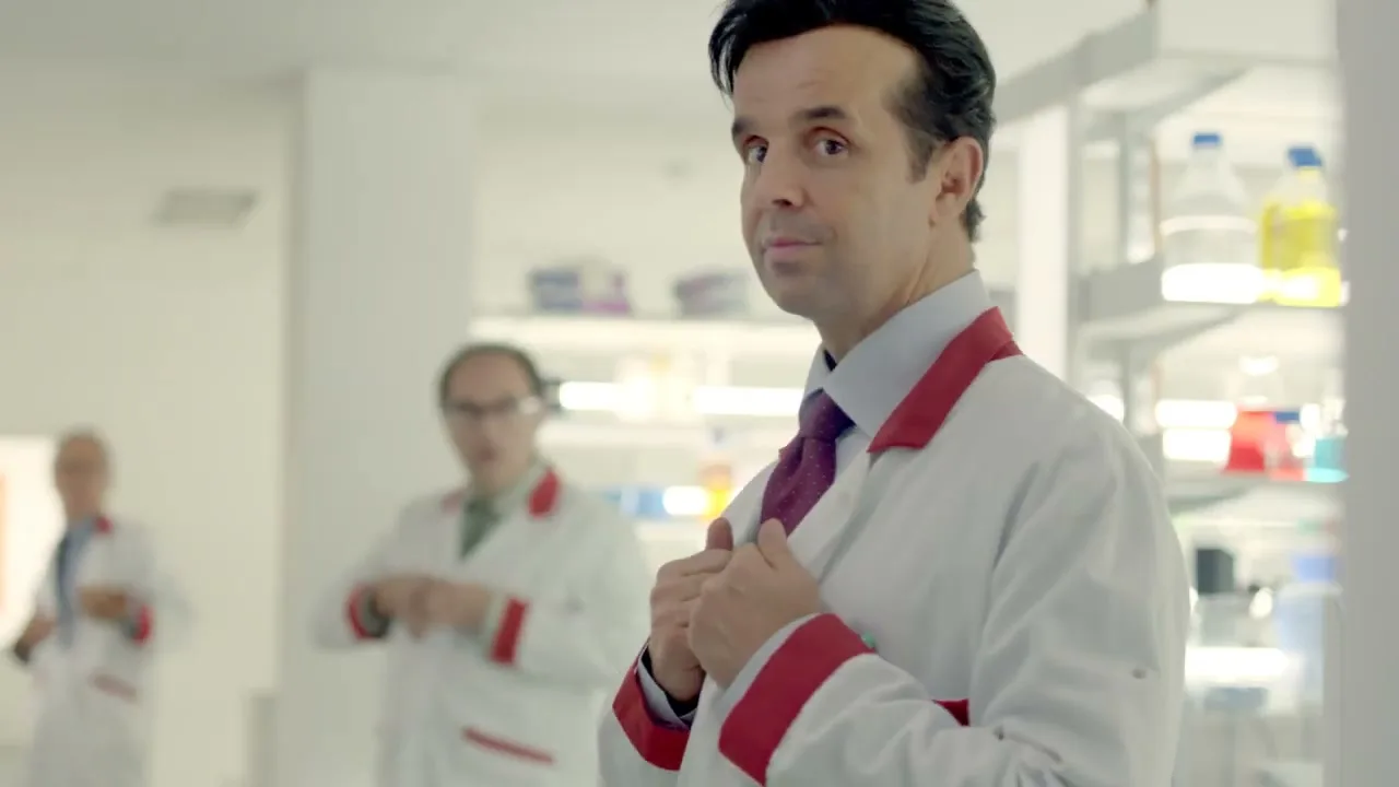 A male doctor in a white lab coat and purple tie turns to face the camera, holding his lapels, in a brightly lit lab.
