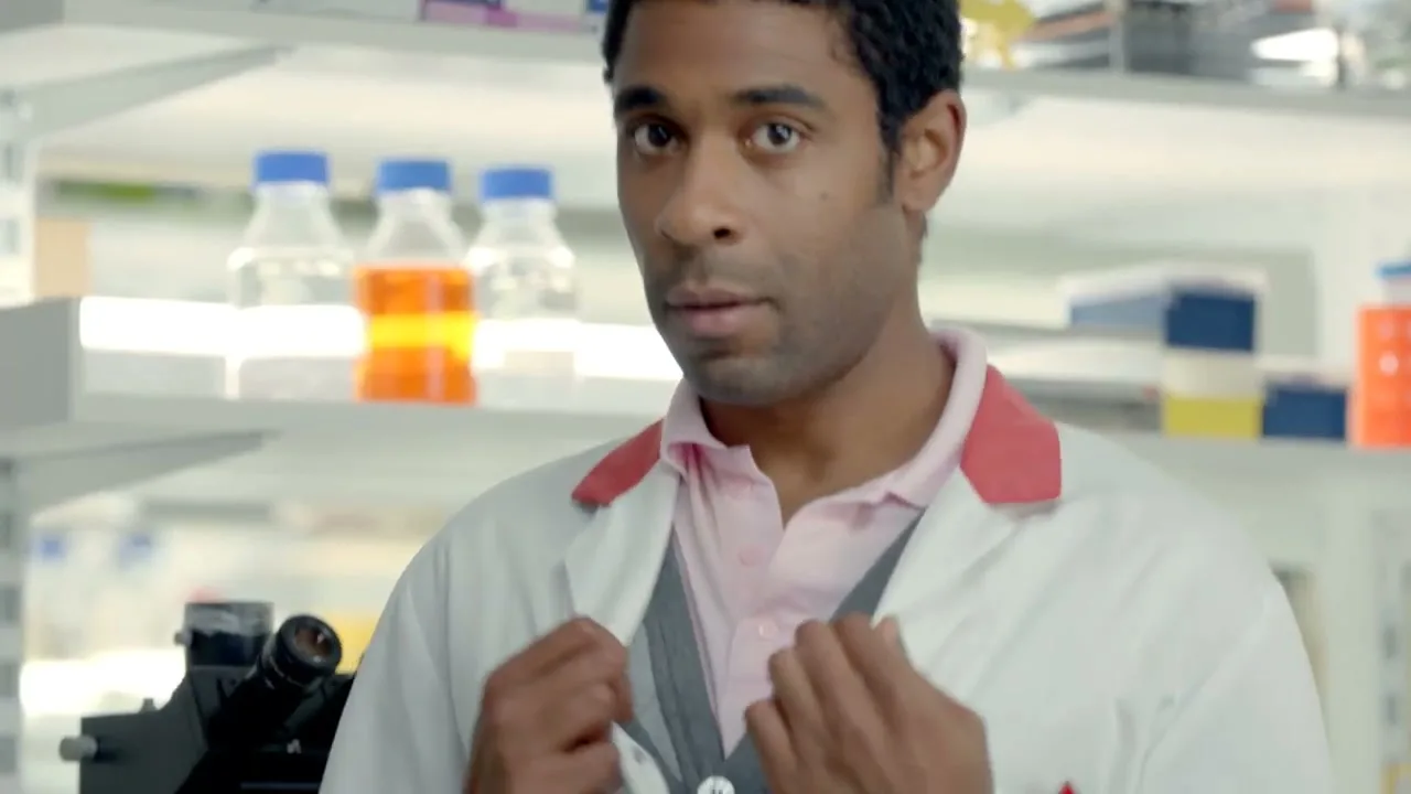 A male scientist in a lab coat and pink polo shirt looks at the camera while unbuttoning his cardigan in a bright laboratory.