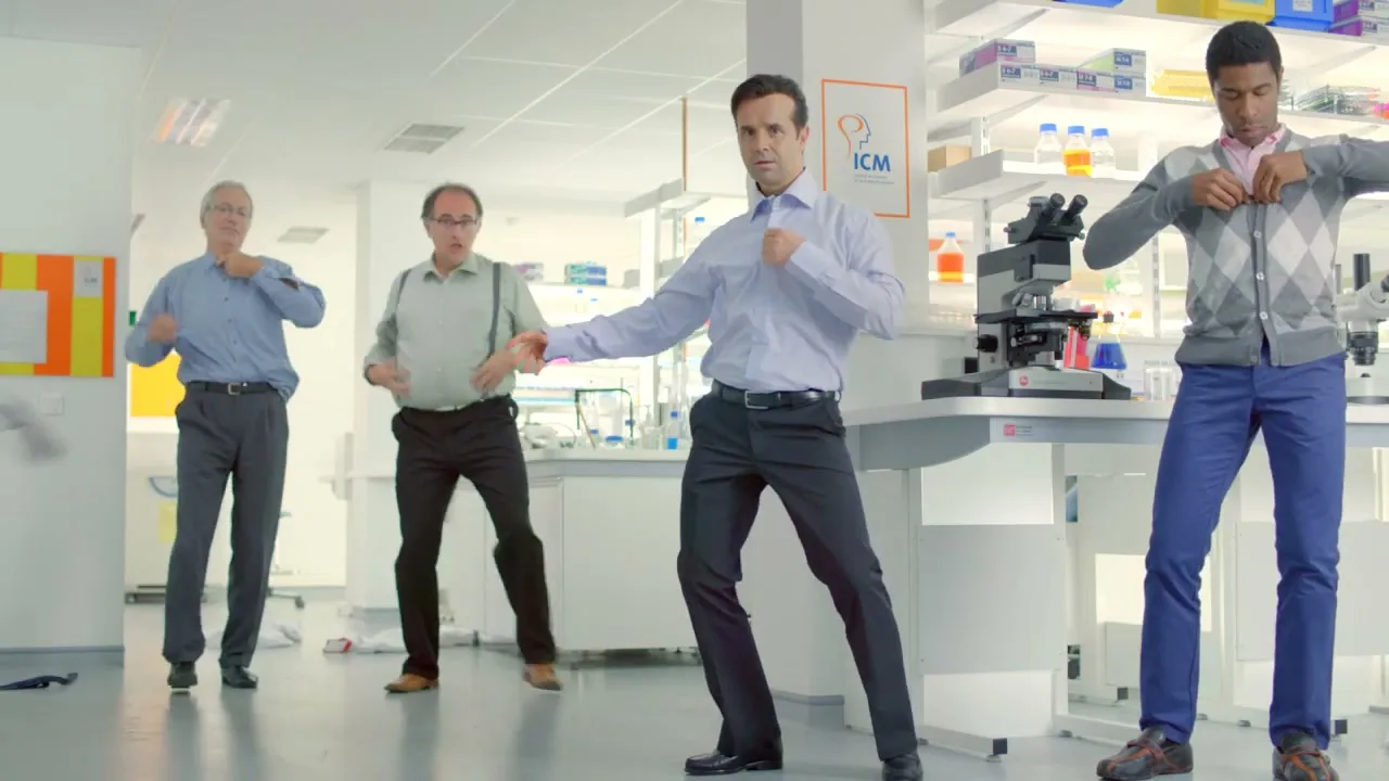 Four male scientists of varying ages dance in a laboratory, removing their shirts in a striptease-like fashion.