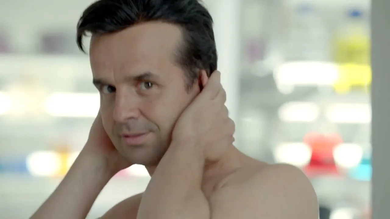 A man with dark hair and bare chest looks at the camera, then looks down and touches his neck, in a bright, blurry background.