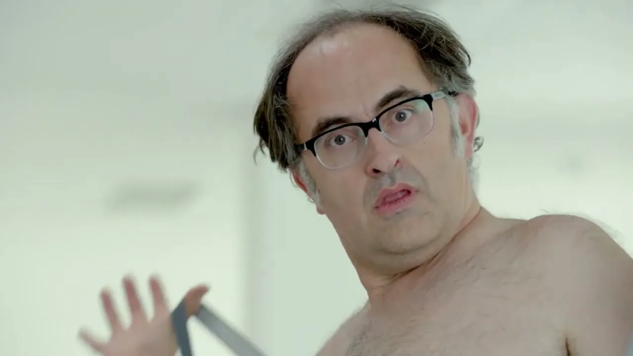 A balding man in his 40s, wearing glasses and suspenders, looks surprised while taking off his shirt in a brightly lit room.