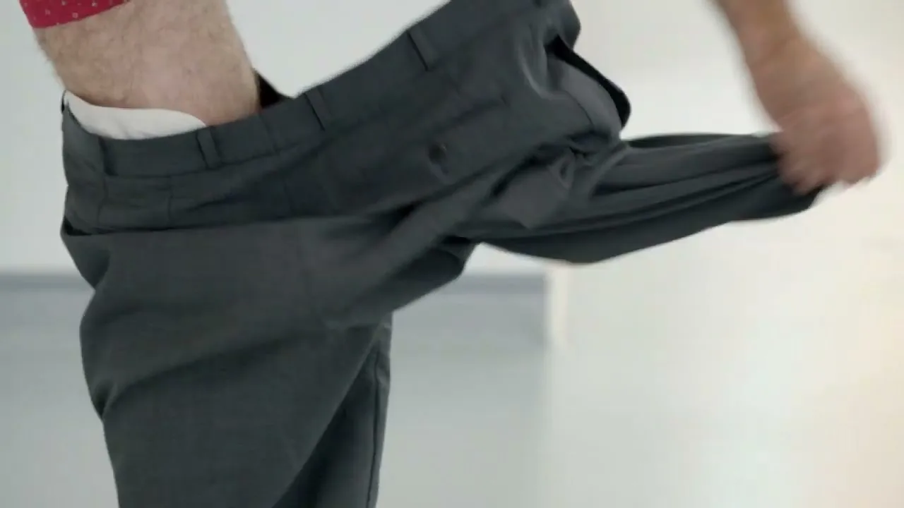 A man in red boxers and grey pants pulls up his pants in a bright, minimalist room.