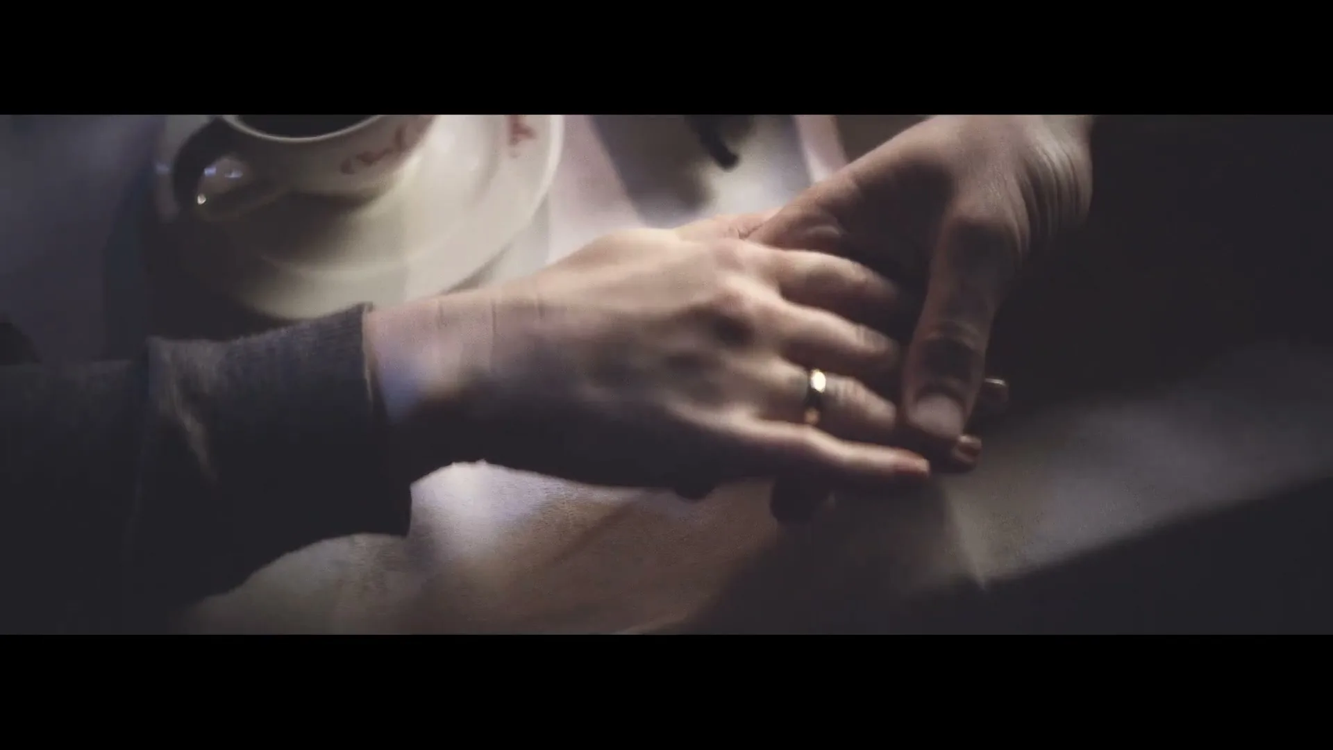 A man and woman hold hands across a table, with a coffee cup visible, in a dimly lit restaurant.