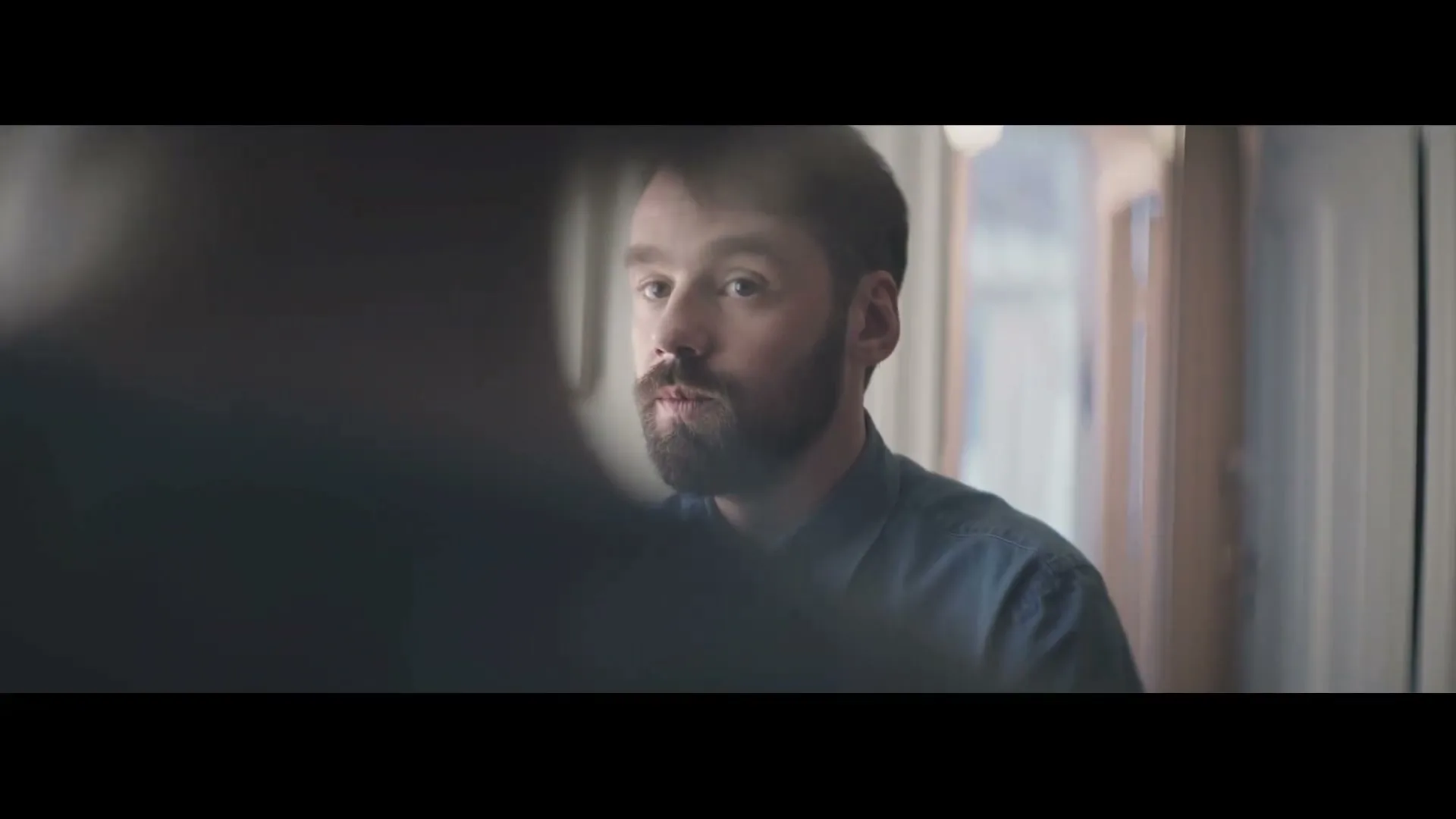 A bearded man in a blue shirt looks intently at someone off-screen, partially obscured by a dark foreground element in an interior setting.
