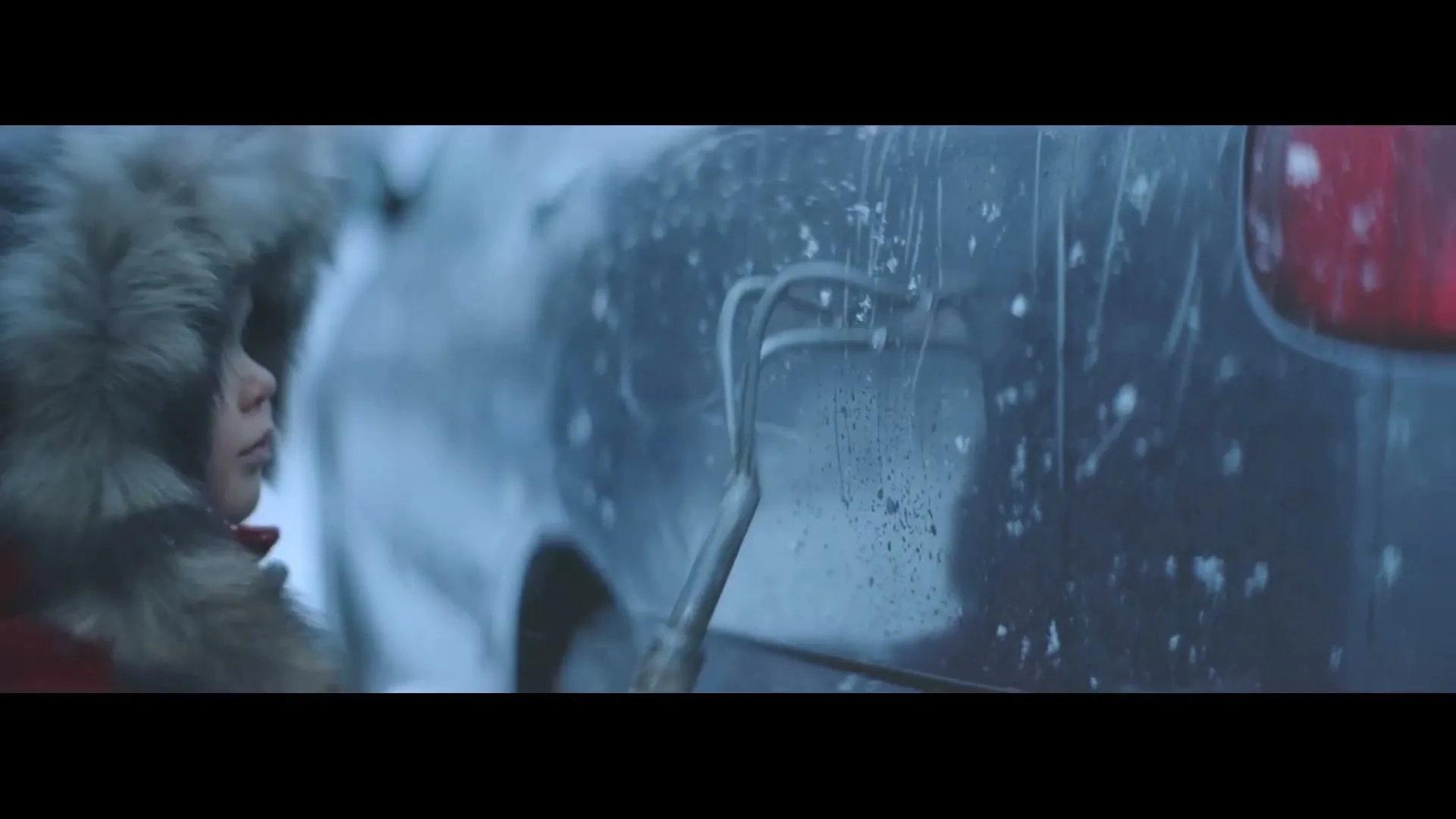 A child in a fur-lined hoody draws a heart in the snow on the side of a dark car in a snowy outdoor setting.