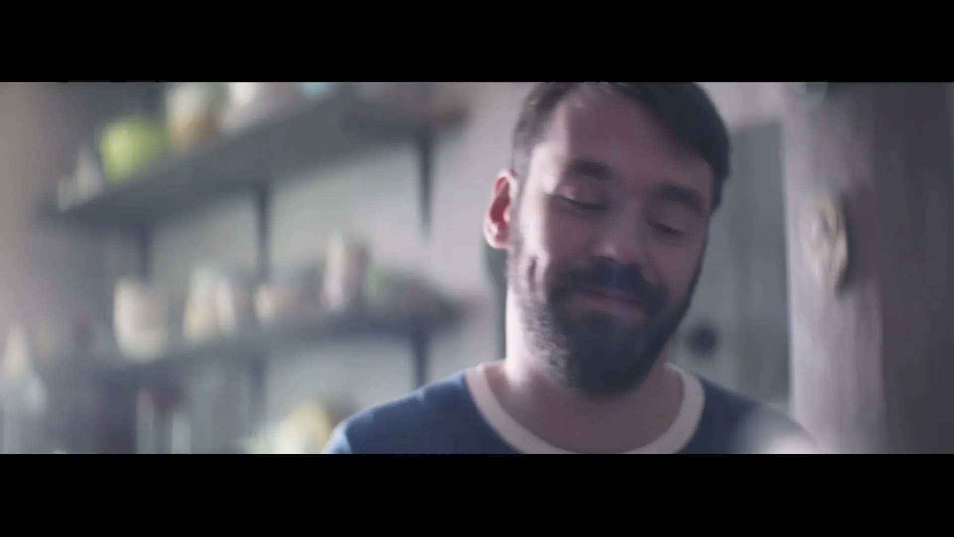 A man with a beard and short dark hair smiles and looks down, wearing a blue and white t-shirt in a kitchen.