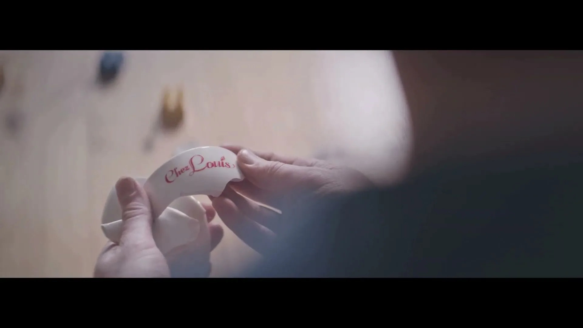 A person's hands carefully piece together a broken white ceramic plate with "Chez Louis" written in red, on a light wooden surface.