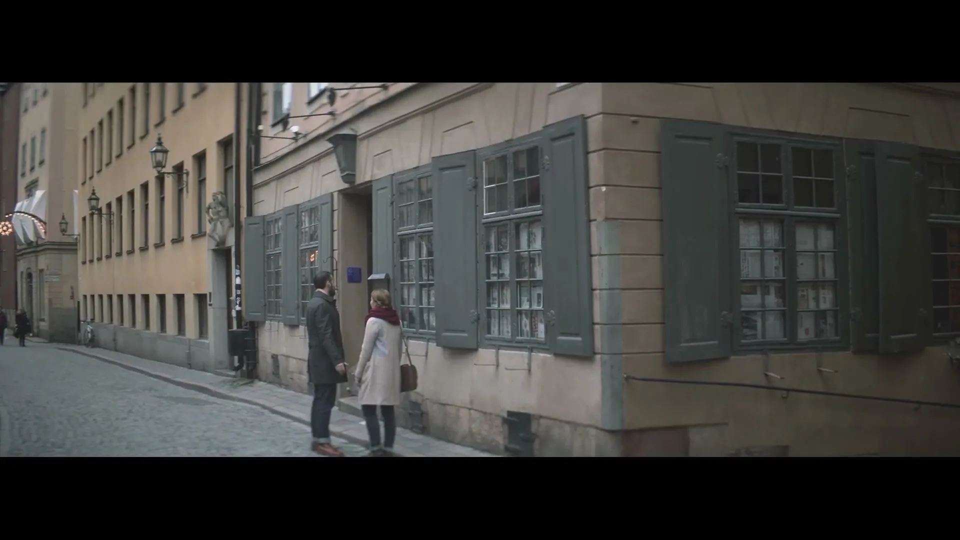 A man and a woman stand on a cobblestone street, looking at each other in front of an old building with green shutters.
