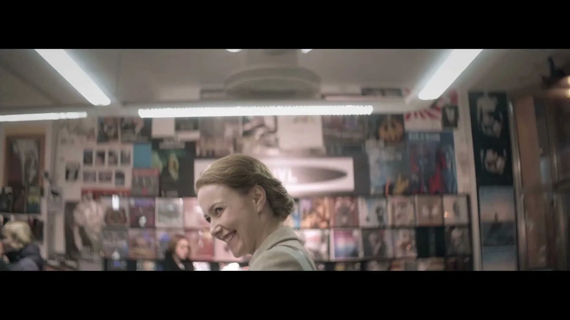 A woman with blonde hair in a bun smiles and looks over her shoulder in a record store, surrounded by album covers.