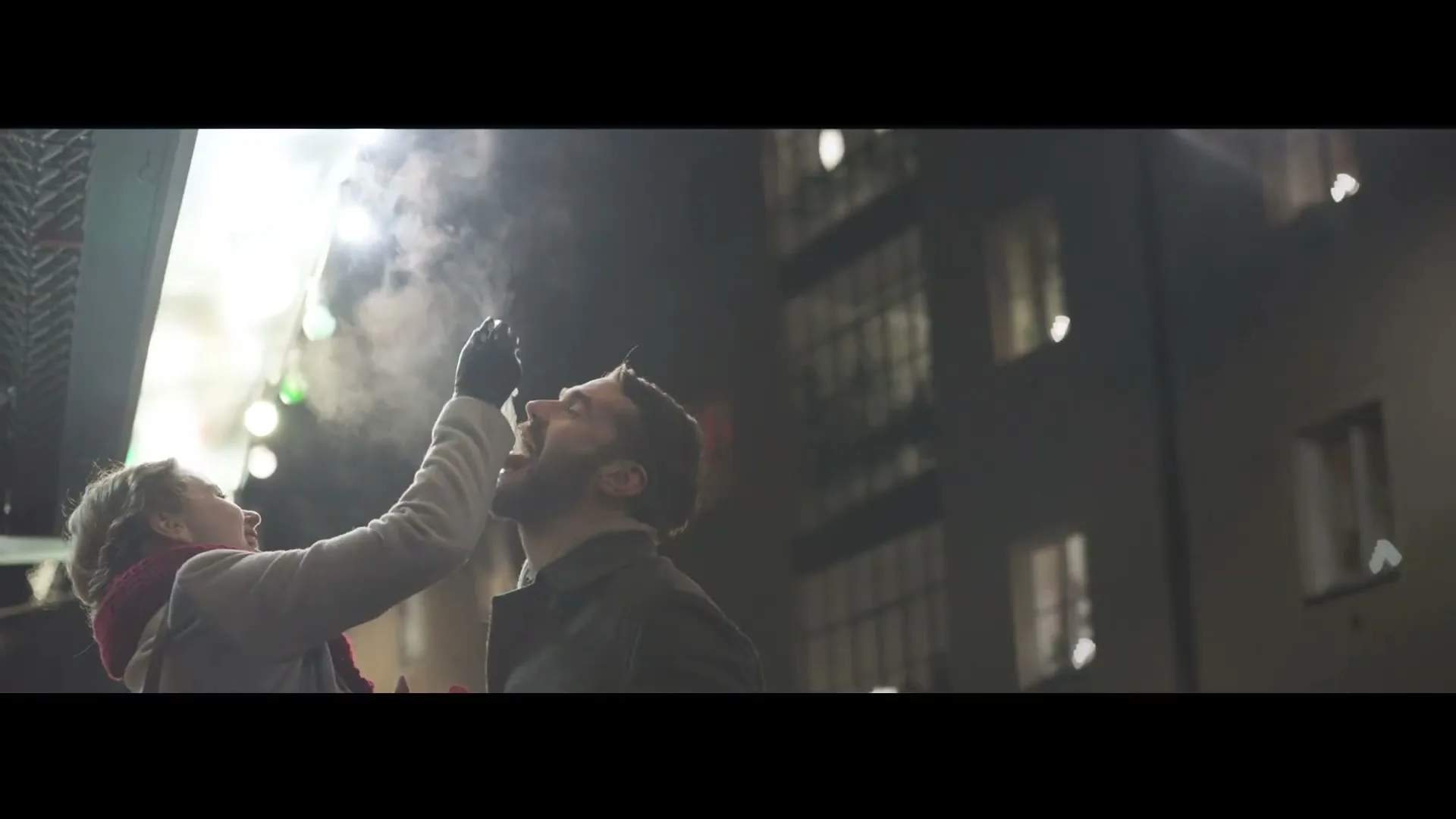 A woman in a coat and scarf sprays whipped cream into a man's open mouth on a city street at night.