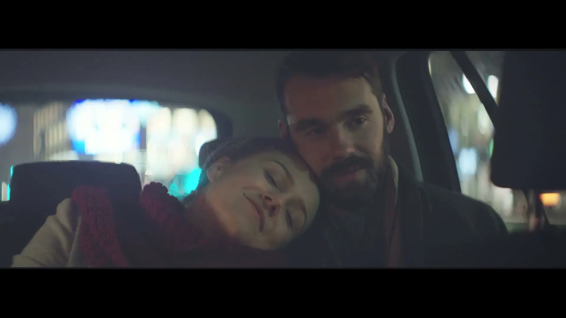 A young woman with a red scarf rests her head on a bearded man's shoulder in the back of a car at night, looking content.