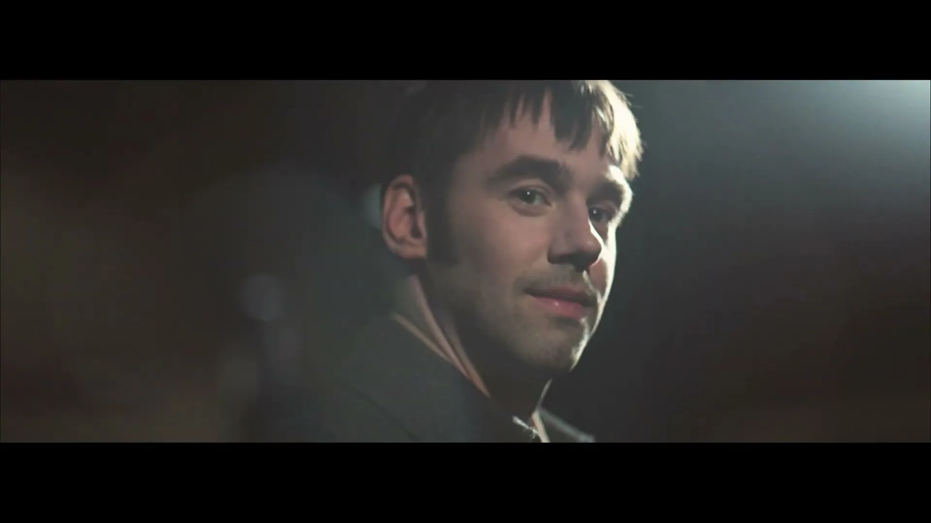 A young man with short hair and a dark coat looks over his shoulder and smiles subtly in a dimly lit interior.