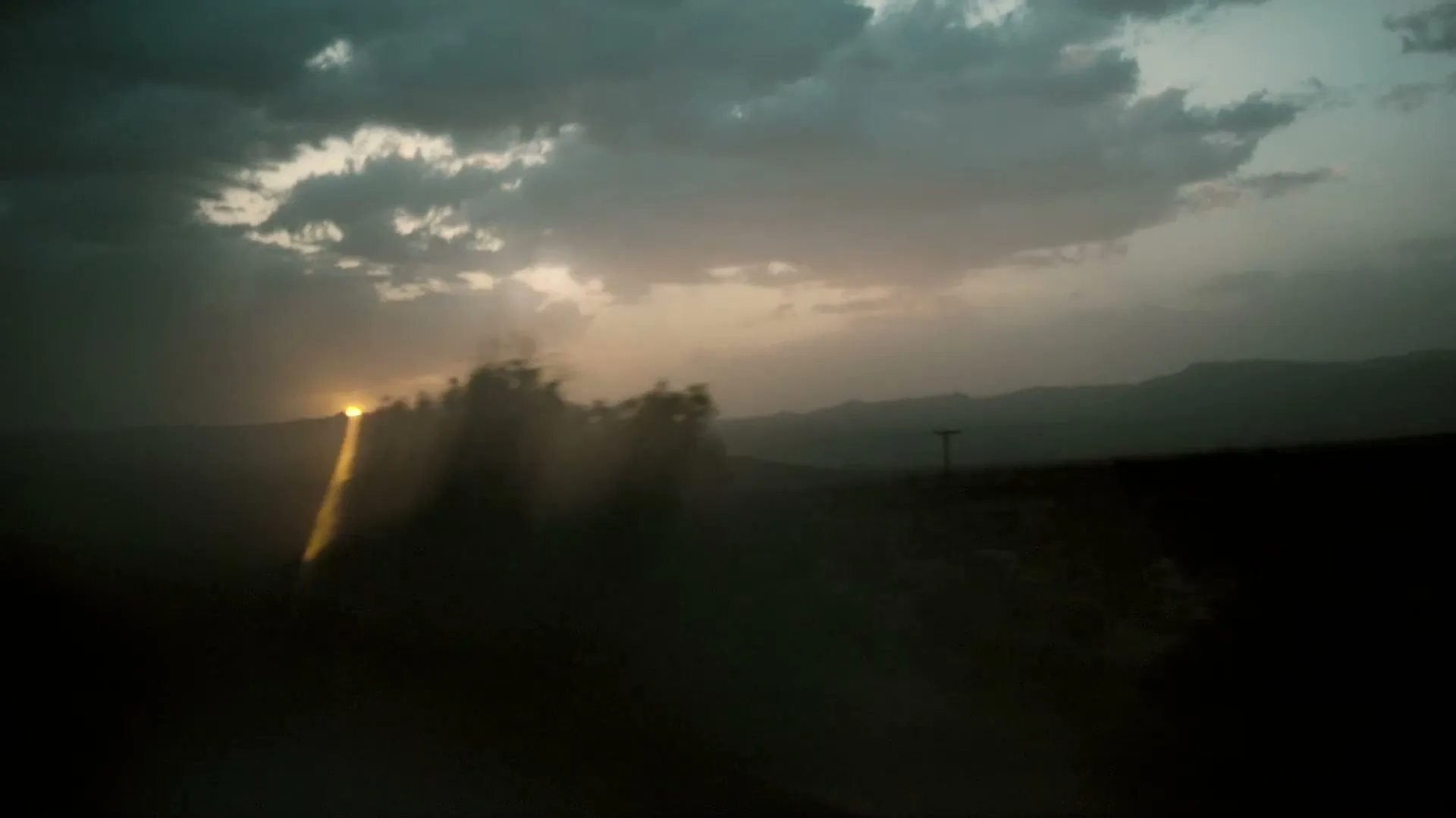 The sun sets behind a distant mountain range, casting a golden glow through dark clouds over a vast, empty landscape.
