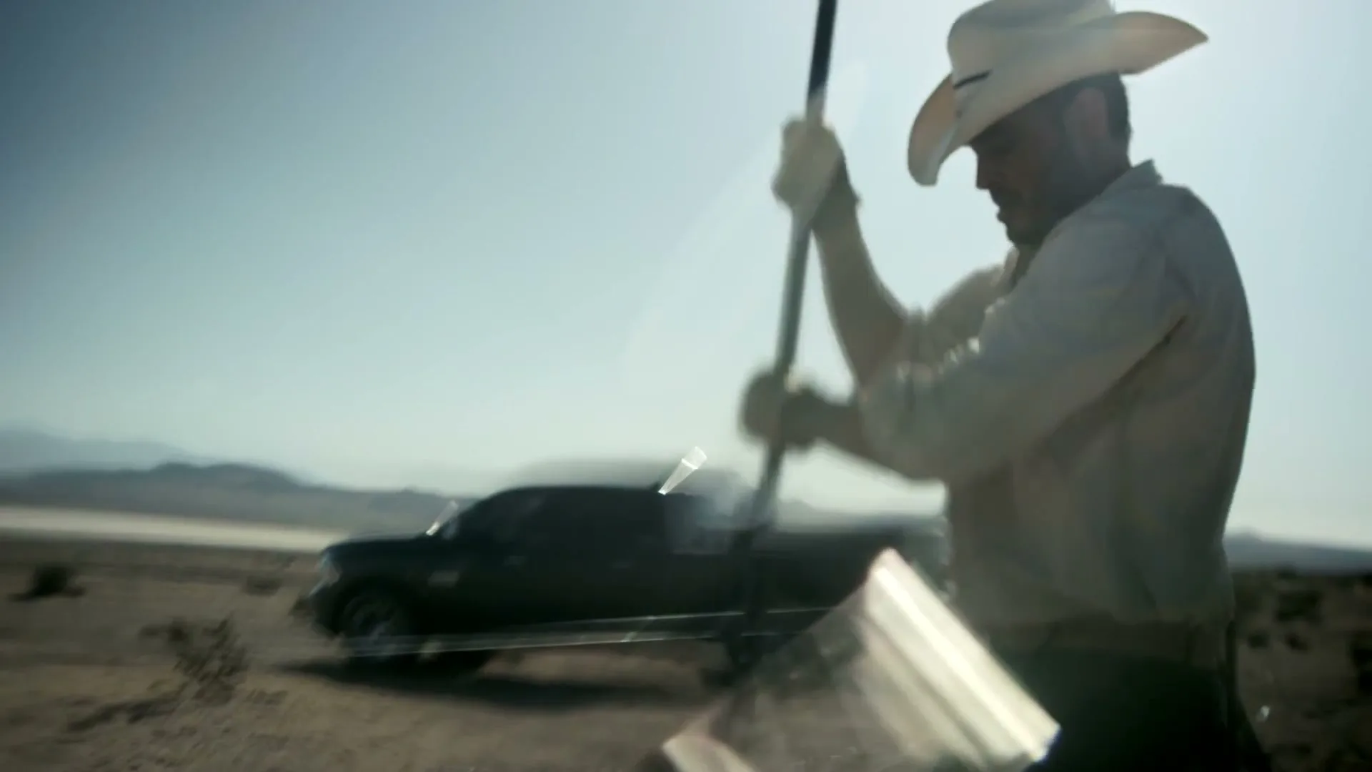 A male cowboy in a white shirt and hat works with a shovel in a desert landscape, with a dark truck in the background under a bright sky.
