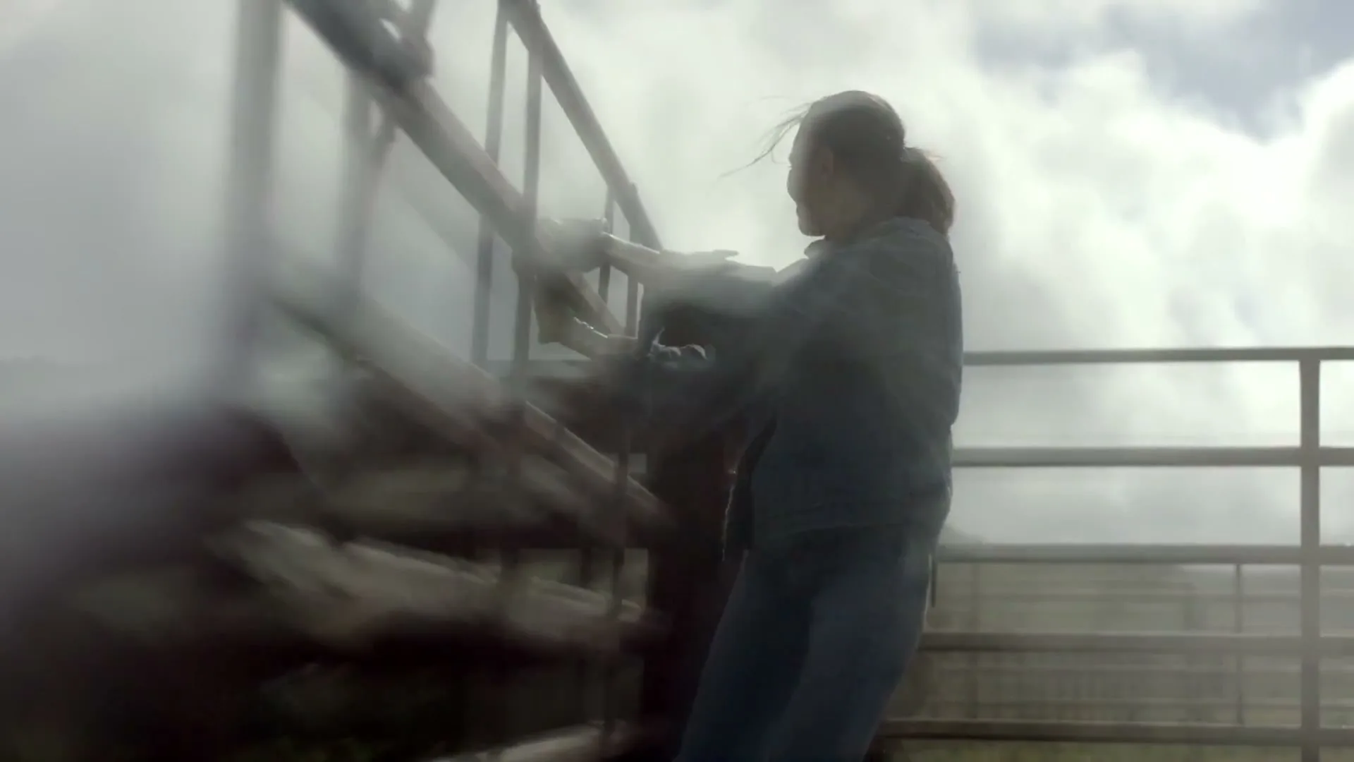 A female rancher in a denim shirt and jeans struggles to close a metal gate on a cloudy day at a ranch.