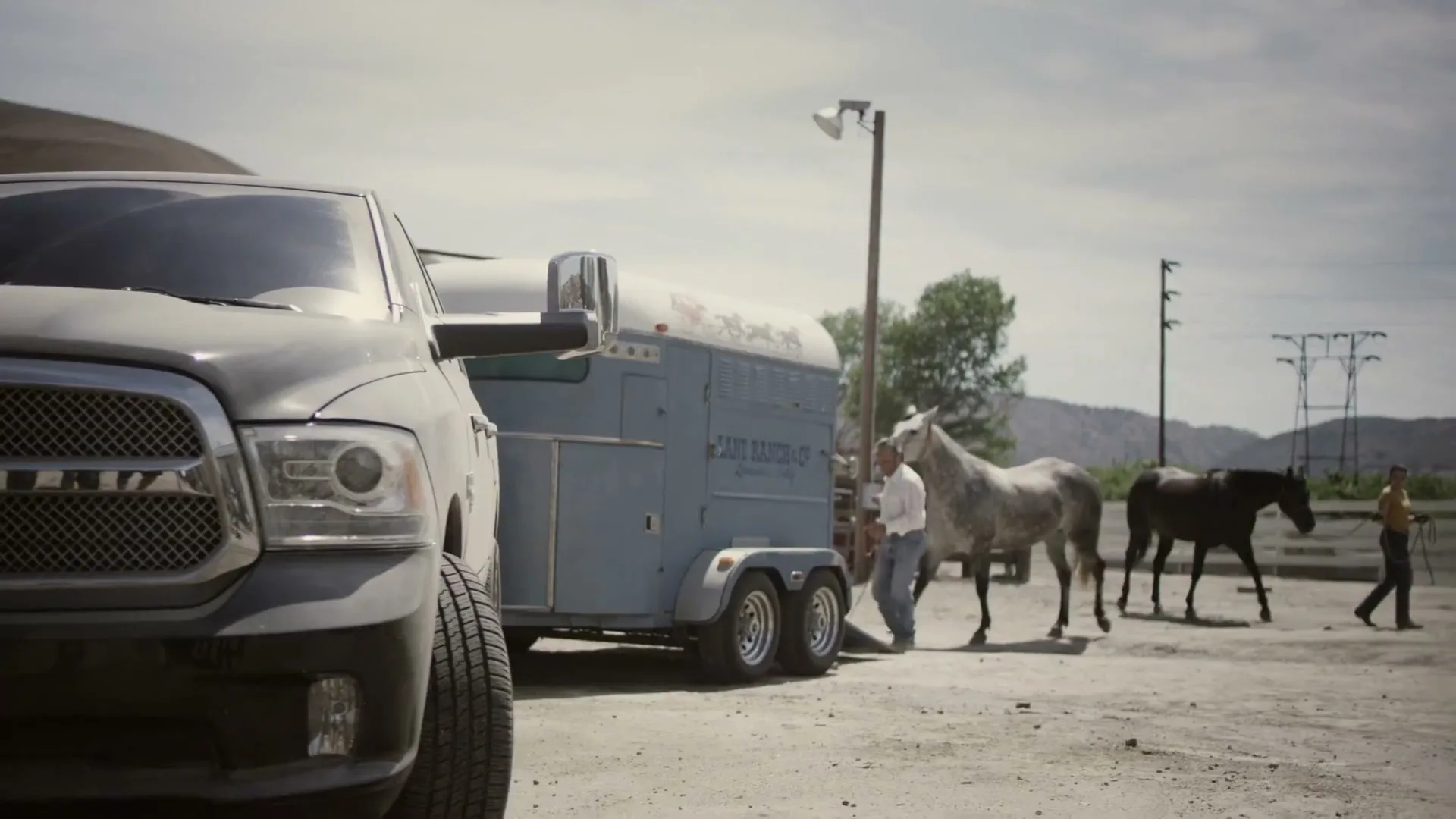 A gray Ram truck is parked next to a horse trailer, while a man and woman lead two horses in a dusty, rural setting.