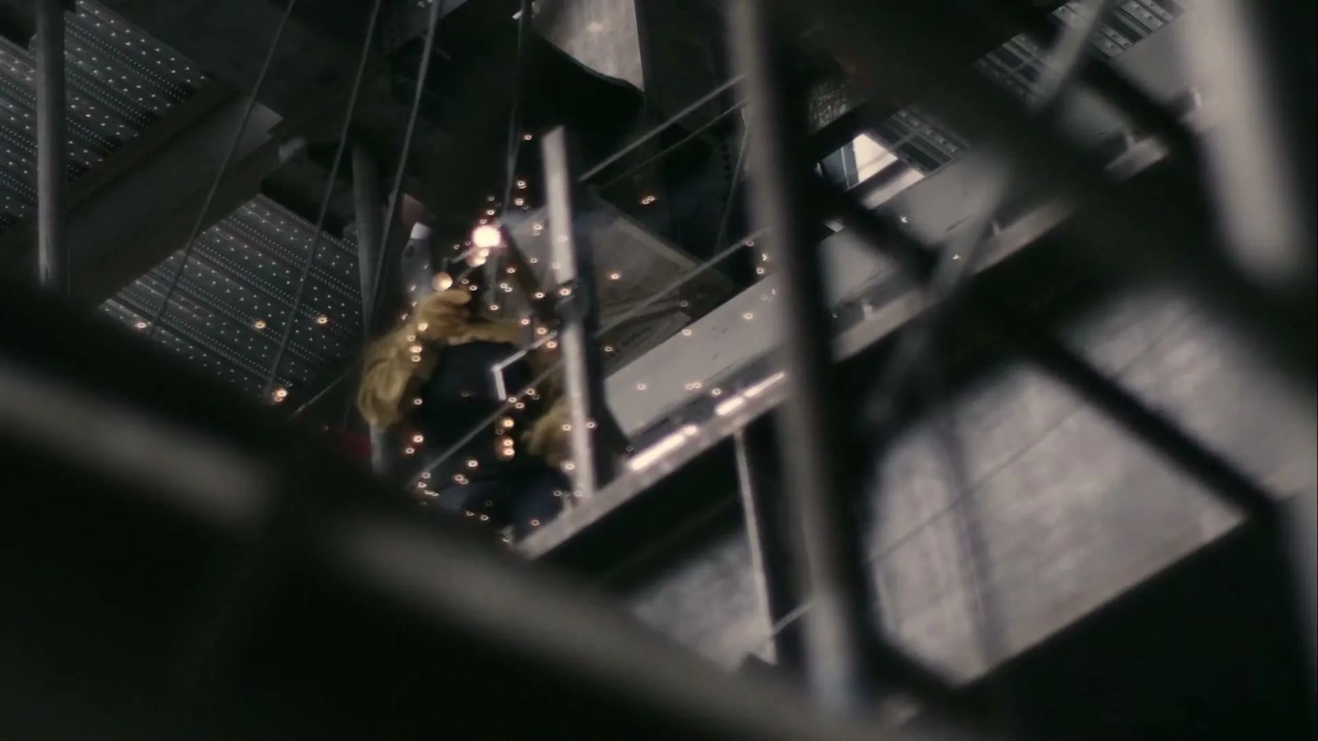 A welder in a yellow jacket and dark pants works on a metal structure, sparks flying, in an industrial setting.