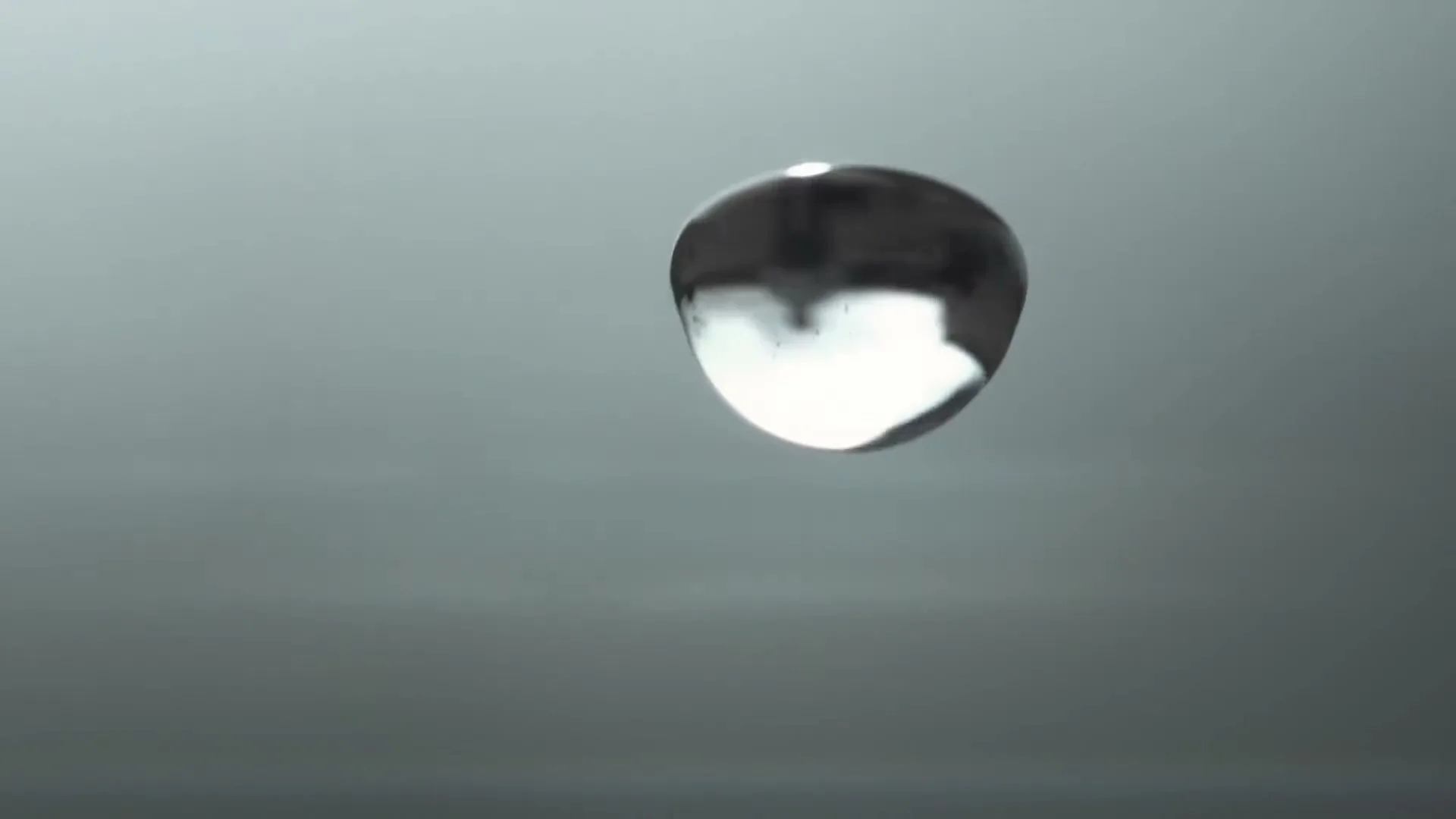 A single, perfectly spherical drop of liquid, reflecting light, floats in mid-air against a plain grey background.