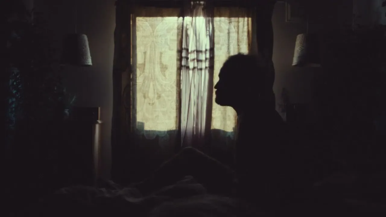 A man sits up in bed, silhouetted against a bright window with patterned curtains in a dark bedroom.
