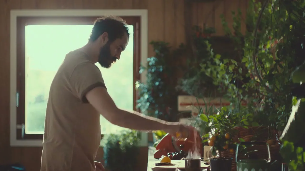 A man with a beard in a t-shirt squeezes a lemon into a steaming cup on a table full of plants in a rustic room.