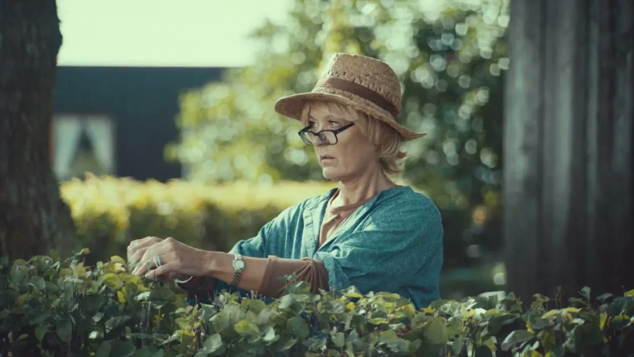 A middle-aged woman in a straw hat and glasses trims a hedge in a suburban garden on a sunny day.
