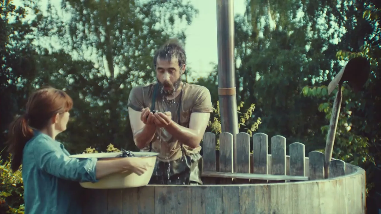A woman in a denim shirt holds a bowl of grapes as a man with a beard and wet hair spits water into a wooden barrel in a garden.