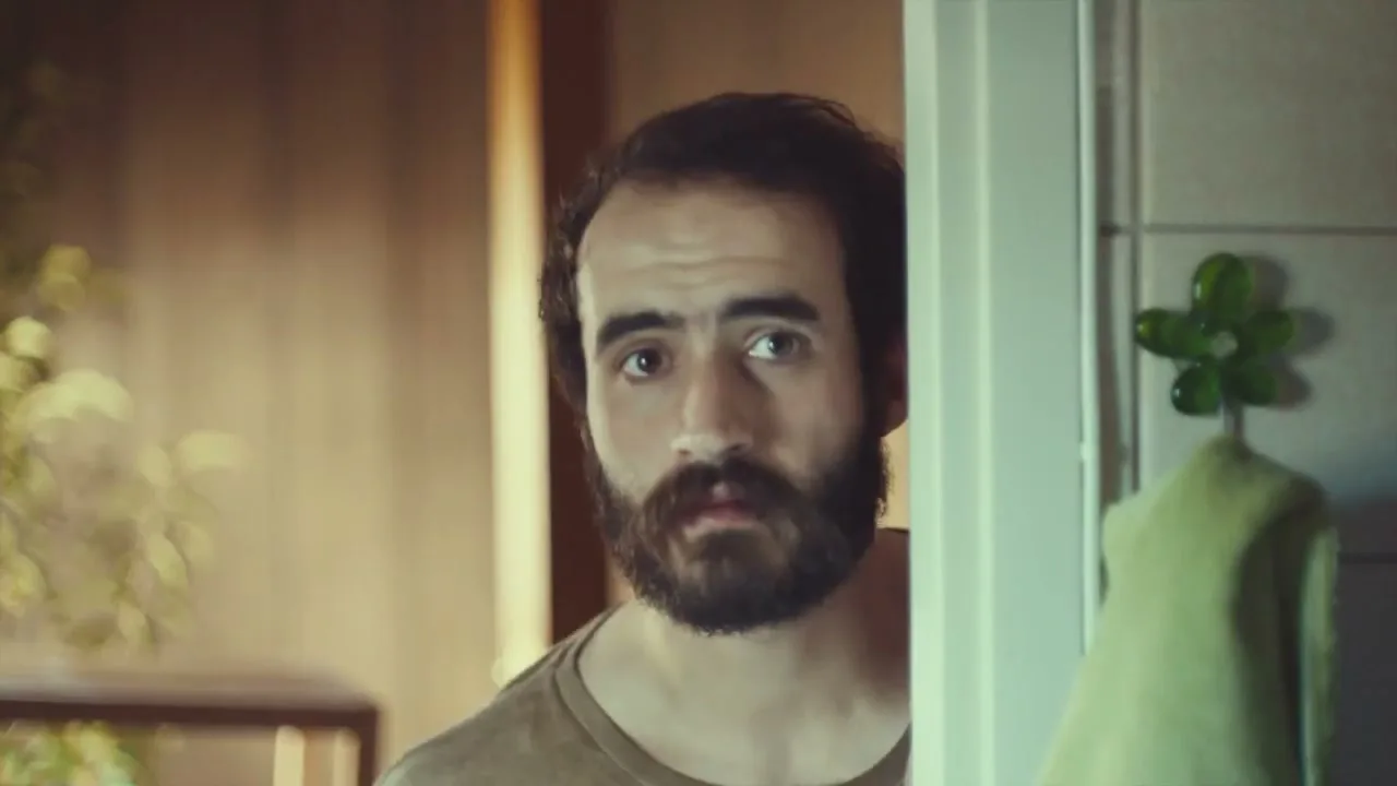 A man with a beard peeks around a doorframe, looking directly at the camera with a curious expression in an interior setting.