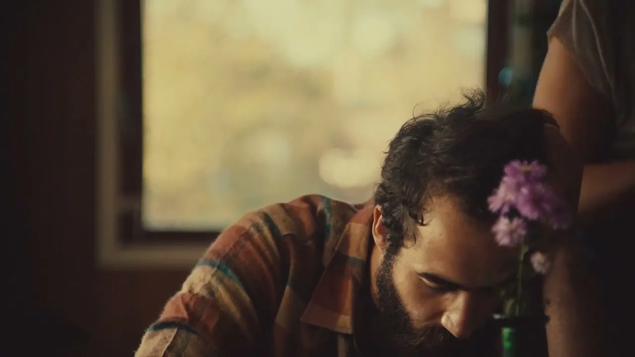 A man with a beard and plaid shirt looks down, then up, as a woman's arm places purple flowers next to him in an interior setting.