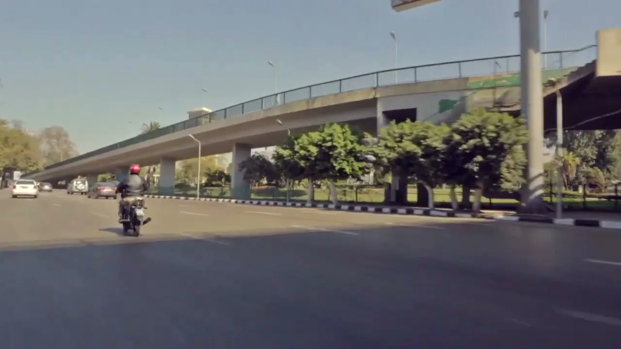 A person on a motorcycle drives away from the camera on a multi-lane road under a highway overpass on a sunny day.