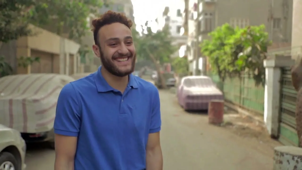 A young man with a beard and short curly hair, wearing a blue polo shirt, smiles and laughs while standing on a residential street with parked cars.