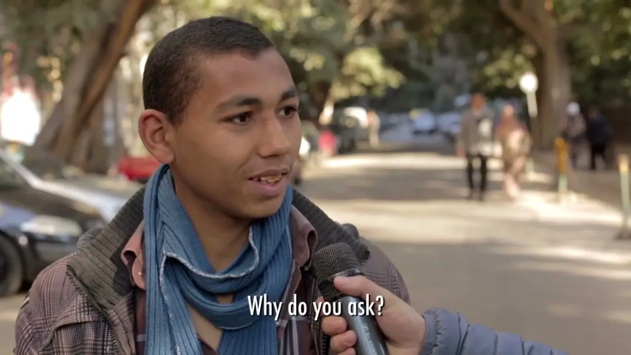 A young man with short hair and a blue scarf is interviewed on a sunny street, holding a microphone and looking thoughtful.