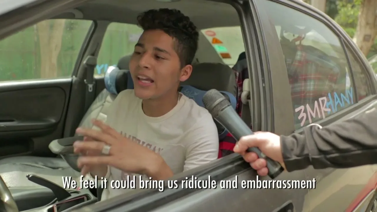 A young man with dark hair and a white long-sleeve shirt sits in a car, gesturing with his hands while being interviewed by someone holding a microphone.