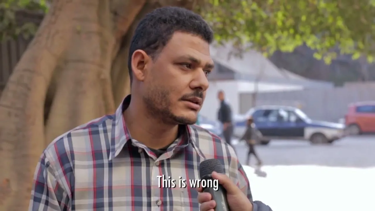 A man in his 30s, wearing a plaid shirt, is interviewed on a street, holding a microphone and looking thoughtful.