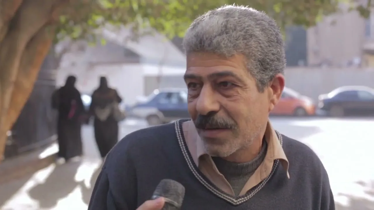 A middle-aged man with a mustache is interviewed on a sunny street, holding a microphone and speaking to an unseen interviewer.