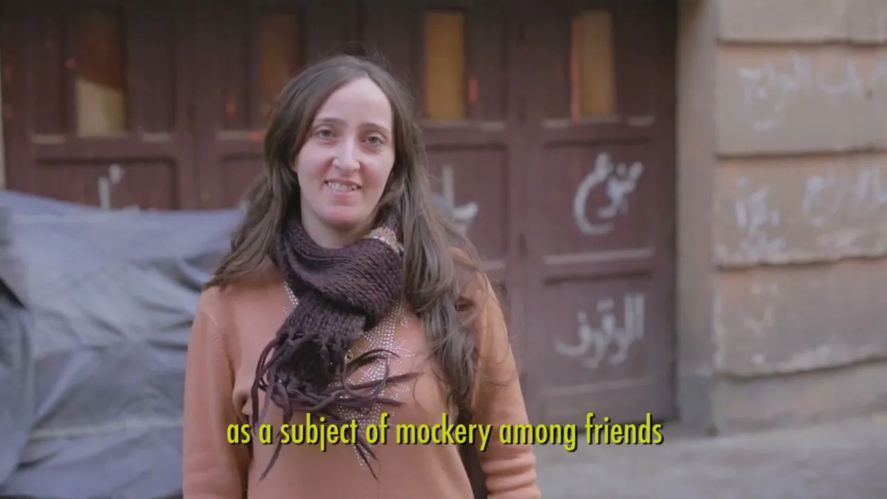 A woman with long brown hair and a brown scarf smiles at the camera, standing in front of a brown door with white Arabic graffiti.