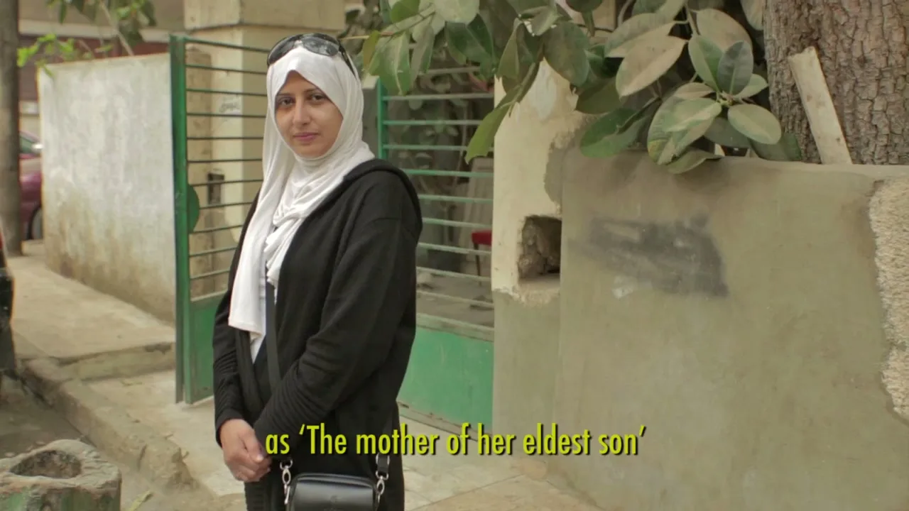 A woman in a white hijab and black hoodie stands on a street, looking at the camera with a neutral expression, next to a wall and green gate.