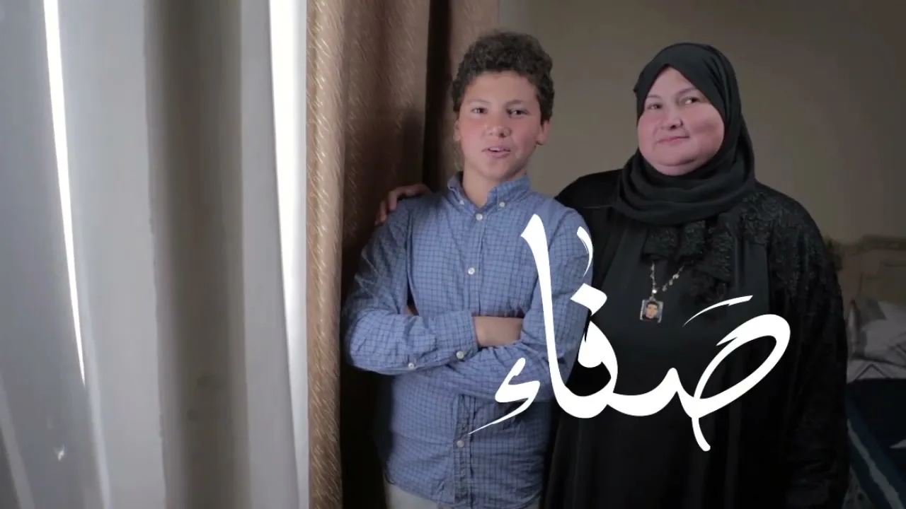 A smiling teen boy in a blue plaid shirt stands with his arm around a smiling older woman in a black hijab and abaya, in a bedroom.