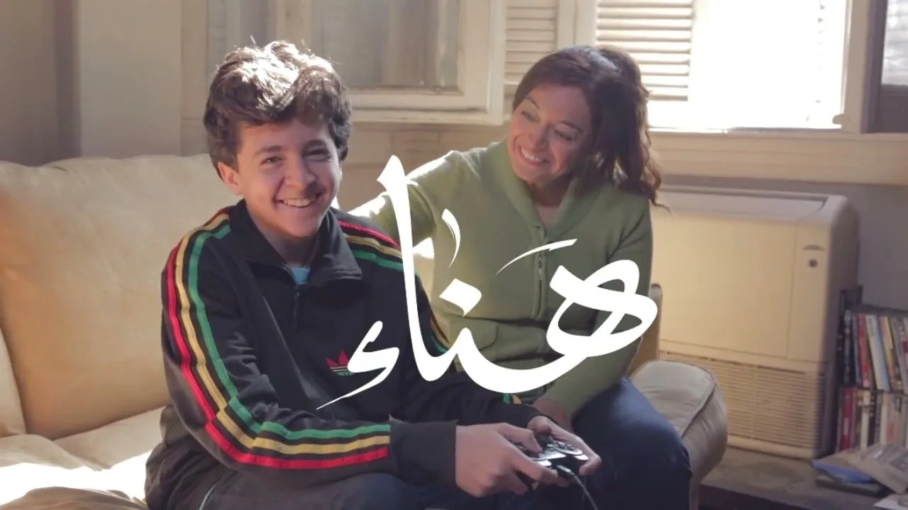 A smiling teen boy in a track jacket plays video games while a woman in a green sweater sits behind him, smiling, in a sunlit living room.