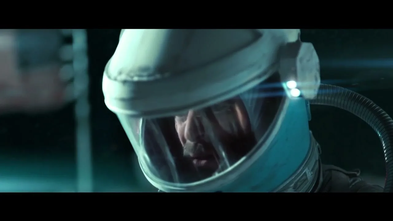 A male astronaut in a white and blue helmet looks down with a serious expression, with a bright pink light and blue lens flare in the background.