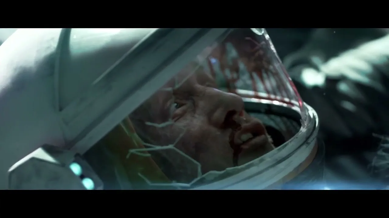 A male astronaut with a bloody face and a cracked helmet is seen in a medium closeup shot, appearing injured and distressed in a dark, possibly space-like environment.