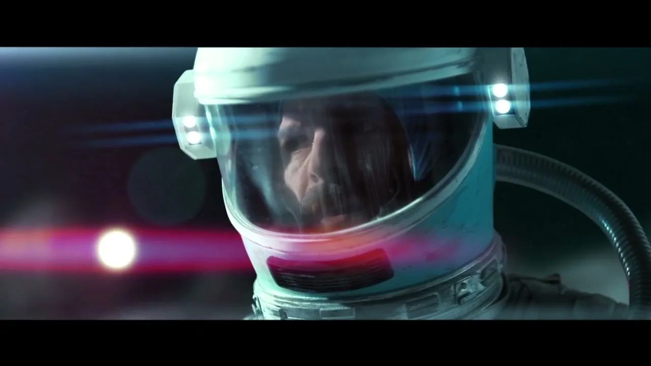 A male astronaut in a white and blue helmet looks around with a serious expression, with a bright light and lens flare in the background.