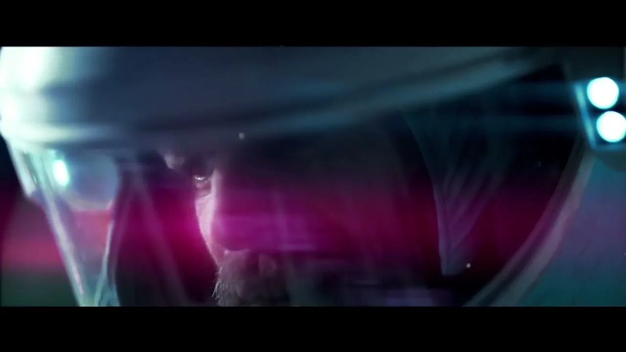 A man in a futuristic astronaut helmet with a beard looks down, with bright pink and blue light flares reflecting on his visor.