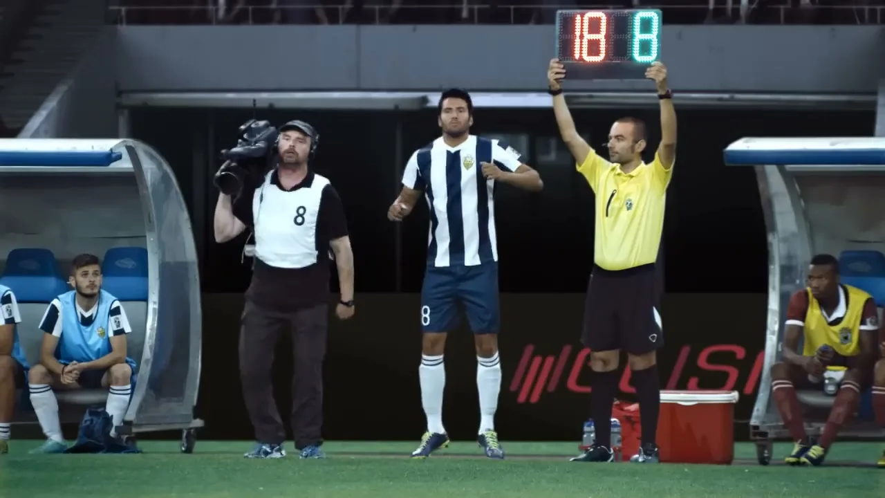 A cameraman, a soccer player, and a referee stand on a soccer field, with the cameraman jumping excitedly.